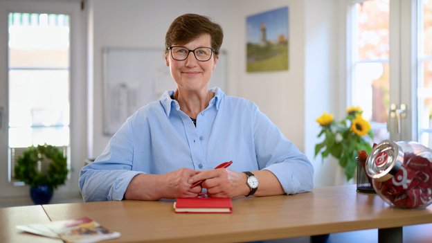 Dörte Kumschließ steht mit hellblauem Hemd an einem Holztisch, hält roten Stift und rotes Notizbuch, im Hintergrund Fenster und Pflanze.