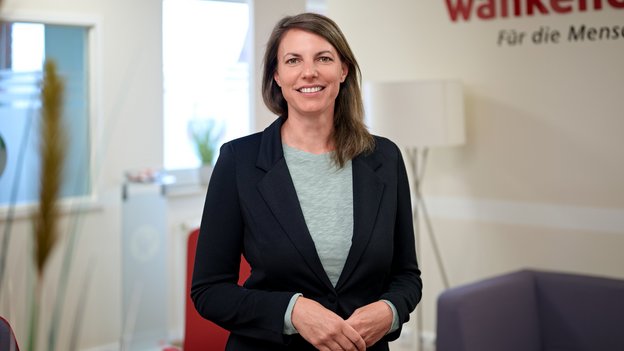 Die Ansprechpartnerin Jana Büll steht in einem Büro und trägt schwarzen Blazer sowie graues Shirt, Hände vor dem Bauch verschränkt.