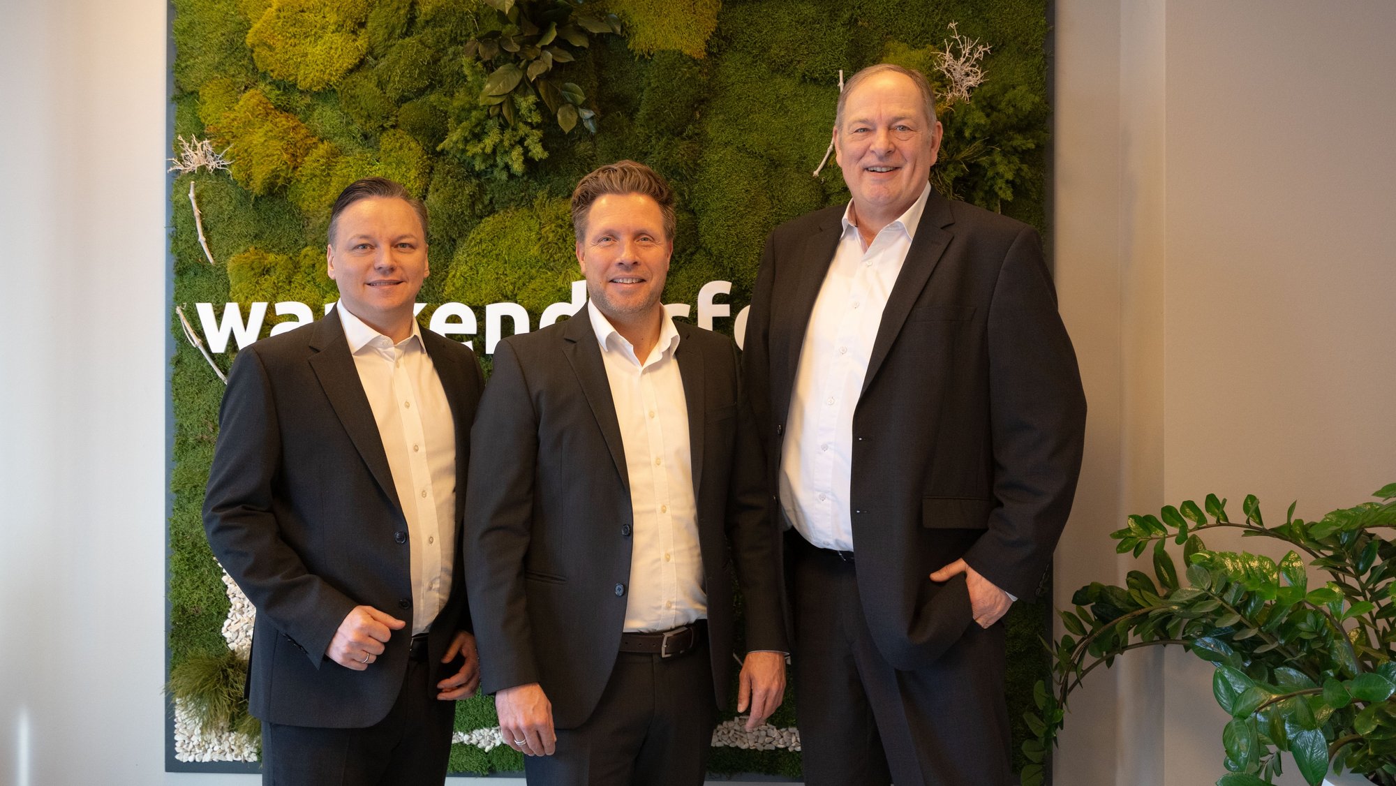 Drei Männer in Anzügen stehen lächelnd vor einer grünen Wand mit Moos und Pflanzen. Sie posieren für ein Gruppenfoto in einer Büroumgebung.