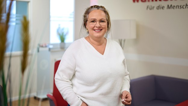 Die Ansprechpartnerin Jessica von Dach steht in einem hellen Raum, trägt einen weißen Pullover und blaue Jeans, eine Hand in der Hosentasche.