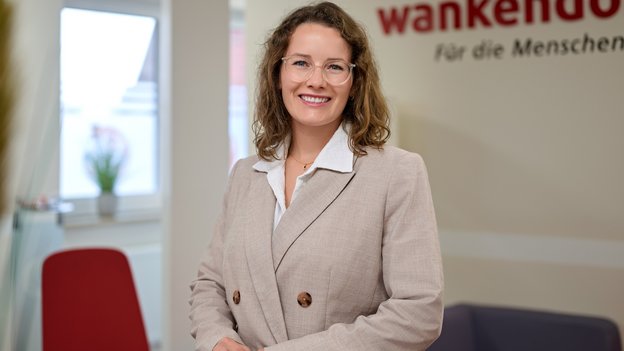 Die Ansprechpartnerin Lisa Sorgenfrei-Koch trägt einen hellbeigen Blazer und eine weiße Bluse, steht vor Wand mit rotem Schriftzug 'wankendorfer Für die Menschen'.