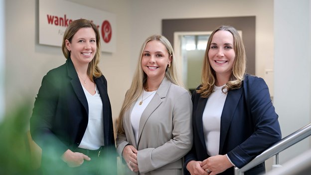 Bente Kiesbye, Marie Kerstan, Fiona Berge in Businesskleidung stehen nebeneinander in einem Büroflur vor einer Wand mit dem Logo der Wankendorfer.